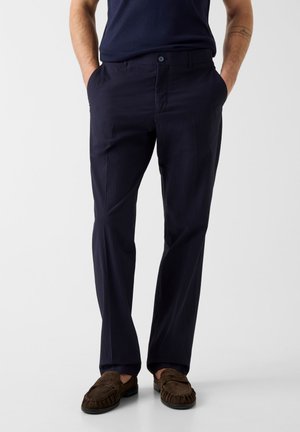 Mężczyzna w ciemnoniebieskich spodniach z rękami w kieszeniach i brązowych mokasynach z zamszu, stoi na tle jasnego, jednolitego tła.