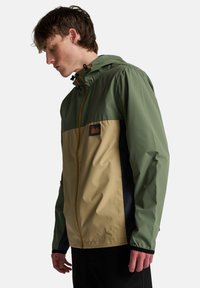 Nepremokavá bunda so zeleným vrchným dielom, béžovou strednou časťou a námorníckymi bokmi. Obsahuje kapucňu a viditeľný zips s oranžovými detailmi.