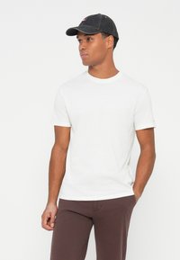 Vīrietis valkā tumši pelēku beisbola cepuri, baltu īsu piedurkņu T-kreklu un brūnas bikses, stāvot ar vienu roku kabatā uz baltas fona.