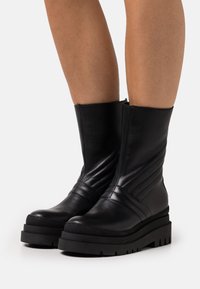 Černé kožené kotníkové boty s vertikálním žebrovaným designem, silnou černou podrážkou a zaoblenou špičkou. Zapínání na zip na vnitřní straně boty.