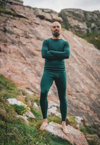 Groene thermische basislaagenset gemaakt van zacht, rekbaar materiaal, bestaande uit een shirt met lange mouwen en aansluitende broek. Model staat op blote voeten op rotsen.