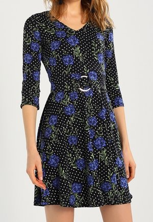 Robe noire à pois blancs et imprimé floral bleu, manches trois-quarts, taille cintrée avec ceinture.