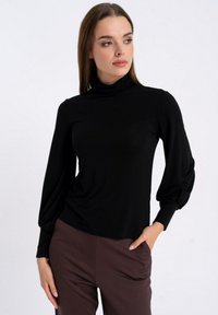 Czarna bluzka z długim rękawem i wysokim kołnierzem, wykonana z teksturowanej tkaniny, z marszczonymi bufkami na rękawach, zestawiona z brązowymi spodniami.