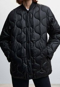 Kurtka pikowana z czarnego błyszczącego materiału, z okrągłym przeszyciem, zapięciem na napy oraz prążkowanym kołnierzem.