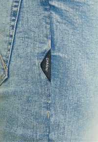 Tissu en denim bleu clair avec une petite étiquette en tissu noir portant l'inscription "CHASIN" cousue dans la couture d'une poche.