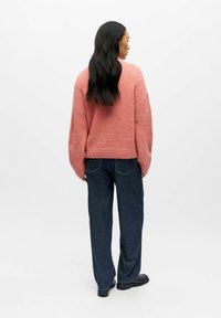 Per didelį rožinį megztinį su nuleistais pečiais, ribotu apačia ir plačiomis rankovėmis, derinamas su tamsiai mėlynais tiesiais džinsais ir juodais batais.