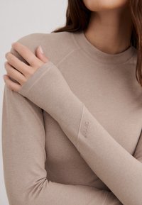 Top de manga larga beige claro con cuello alto y puños alargados. Textura suave con un diseño sin costuras y detalle de logo en la manga.