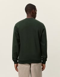 Σκούρο πράσινο φούτερ με στρογγυλή λαιμόκοψη και μακρύ μανίκι. Λείο ύφασμα με ραμμένα μανίκια και στρίφωμα, φορεμένο πάνω από ανοιχτόχρωμο παντελόνι.