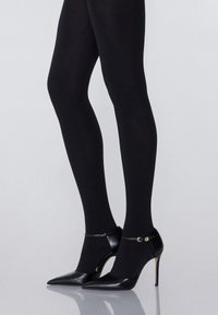 Schwarze High Heels mit spitzer Zehe, Knöchelriemen und goldenen Beschlägen. Getragen mit undurchsichtigen schwarzen Strumpfhosen. Glattes Lederdesign.