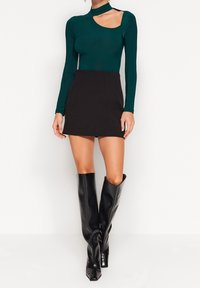 Grüner Bodysuit mit langen Ärmeln und einem Cut-Out-Detail, kombiniert mit einem schwarzen Minirock und knielangen, glänzenden schwarzen Stiefeln.