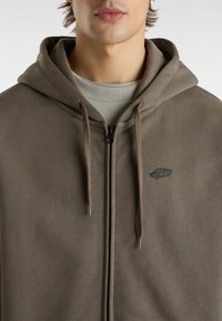 Brązowa bluza z kapturem na zamek, wykonana z miękkiego materiału, z małym logo na klatce piersiowej i sznurkami do ściągania. Prosty, minimalistyczny design.