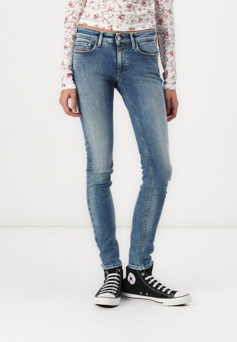 Karcsú, világoskék farmernadrág, fakó mosással, öt zsebbel, enyhe kopással és slim fit szabással. Fekete magas szárú sportcipővel párosítva.