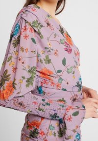 Žena v purpurových květinových šatech s nabíranými rameny a smockovanými manžetami, ukazující horní část těla a ruku opřenou u pasu.