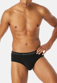 Schwarze Baumwollslips mit einem logoed Bund, die eine enge Passform und eine glatte Textur bieten, auf einem Modell in einer neutralen Umgebung präsentiert.
