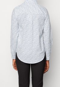 Camicia bianca con un motivo floreale blu, maniche lunghe, colletto abbottonato, orlo curvo e una superficie liscia. Indossata con pantaloni scuri.