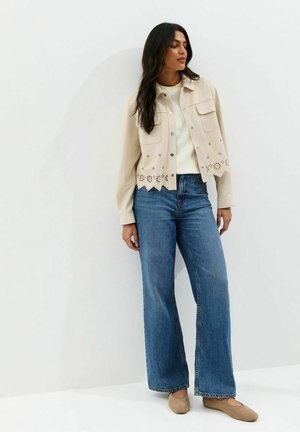 Vrouw met beige jas met bloemenvormige uitsnijdingen, witte trui, wijde blauwe jeans en beige platte schoenen, staand tegen een witte muur.