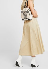 Sac à dos en tissu beige tissé avec des accents noirs, forme courbée et poignées supérieures ; présente un motif damier et une quincaillerie dorée.