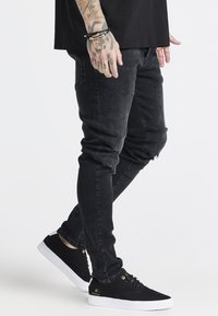 Jeans negros desgastados con un ajuste slim, que presentan detalles rasgados en las rodillas, combinados con zapatillas negras y una camiseta negra sin estampado.