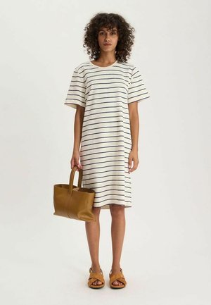 Femme aux cheveux bouclés portant une robe blanche longueur genou avec des rayures horizontales noires, tenant un sac à main marron et portant des sandales marron clair.