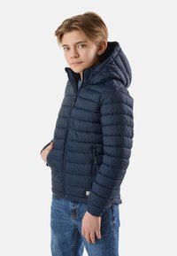 Garçon portant une veste matelassée à capuche bleu marine et un jean bleu, debout avec une main dans la poche de la veste, sur un fond uni.