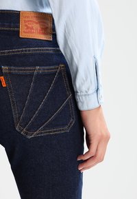 Mörk denimjeans med en distinkt bågad fickdesign, en tan läderlappar på baksidan och en ljusblå långärmad skjorta.