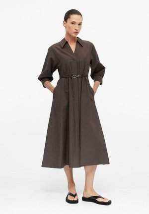 Frau mit dunklen Haaren in einem braunen, langärmeligen Midi-Kleid mit Knöpfen und dünnem Gürtel, Hände in den Taschen, trägt schwarze Plateau-Sandalen.
