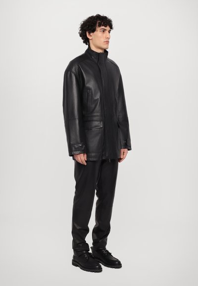 Chaqueta de cuero negra con un alto cuello, que presenta cierre frontal con cremallera, dos bolsillos laterales y una textura suave. Combinada con pantalones negros y calzado.