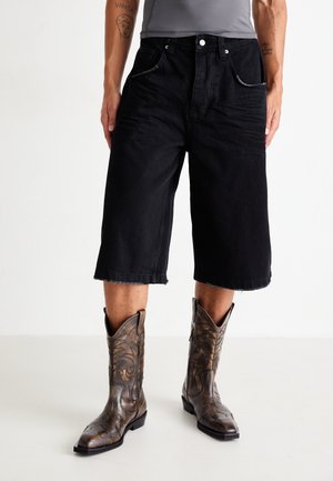 Schwarze Denim-Culottes mit rohem Saum, versehen mit Kontrastnähten und Fronttaschen, kombiniert mit braunen, kunstvoll gestalteten Cowboy-Stiefeln.
