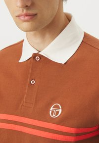 Rudas polo marškinėliai su balta apykakle, mygtukų detalė, atitinkanti rudos spalvos, ir dvi horizontaliomis oranžinėmis juostelėmis su logotipu ant krūtinės.