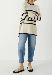Lichtbeige trui met zwarte strepen, geribbelde textuur en lange mouwen, gecombineerd met lichtblauwe denim jeans en beige sandalen met bandjes en zwarte zolen.
