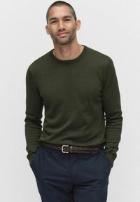 Oliivinvihreä neulepaita pyöreällä kaula-aukolla ja pitkillä hihoilla, yhdistettynä tummansinisisiin housuihin ja punottuun ruskeaan nahkavyöhön.