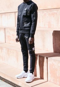 Černá mikina a tepláky s několika kapsami, bílými nápisy a žlutým detailem. Kombinováno s bílými sportovními botami.