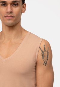 Top beige senza maniche a V, indossato da un modello, con una texture liscia. Un tatuaggio dettagliato è visibile sulla parte superiore del braccio.