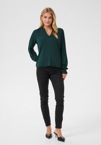 Dunkelgrüne Bluse mit langen Ärmeln, V-Ausschnitt, lockerer Passform und gerüschten Manschetten, kombiniert mit schmalen schwarzen Hosen und spitzen schwarzen Schuhen.