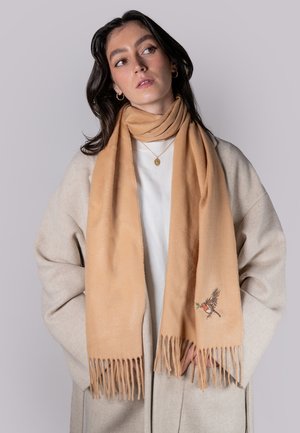 Beige scarf made of soft material, featuring fringe edges and an embroidered bird design. Worn over a light-colored sweater and coat.