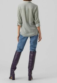 Osoba ubrana w zielono-białą pionową w paski koszulę z podwiniętymi rękawami, niebieskie dżinsy i fioletowe buty do kolan, stojąca plecami.