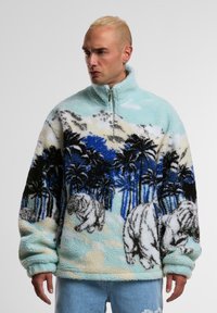 Giacca fleece zip-up blu chiaro e soffice, decorata con tigri bianche e palme in nero e blu, caratterizzata da una superficie morbida e testurizzata.
