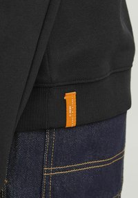 Svart sweatshirt med texturerad tyg och ribbad nederkant. En orange etikett är fäst på sidan med vit text, i kombination med mörk denim.