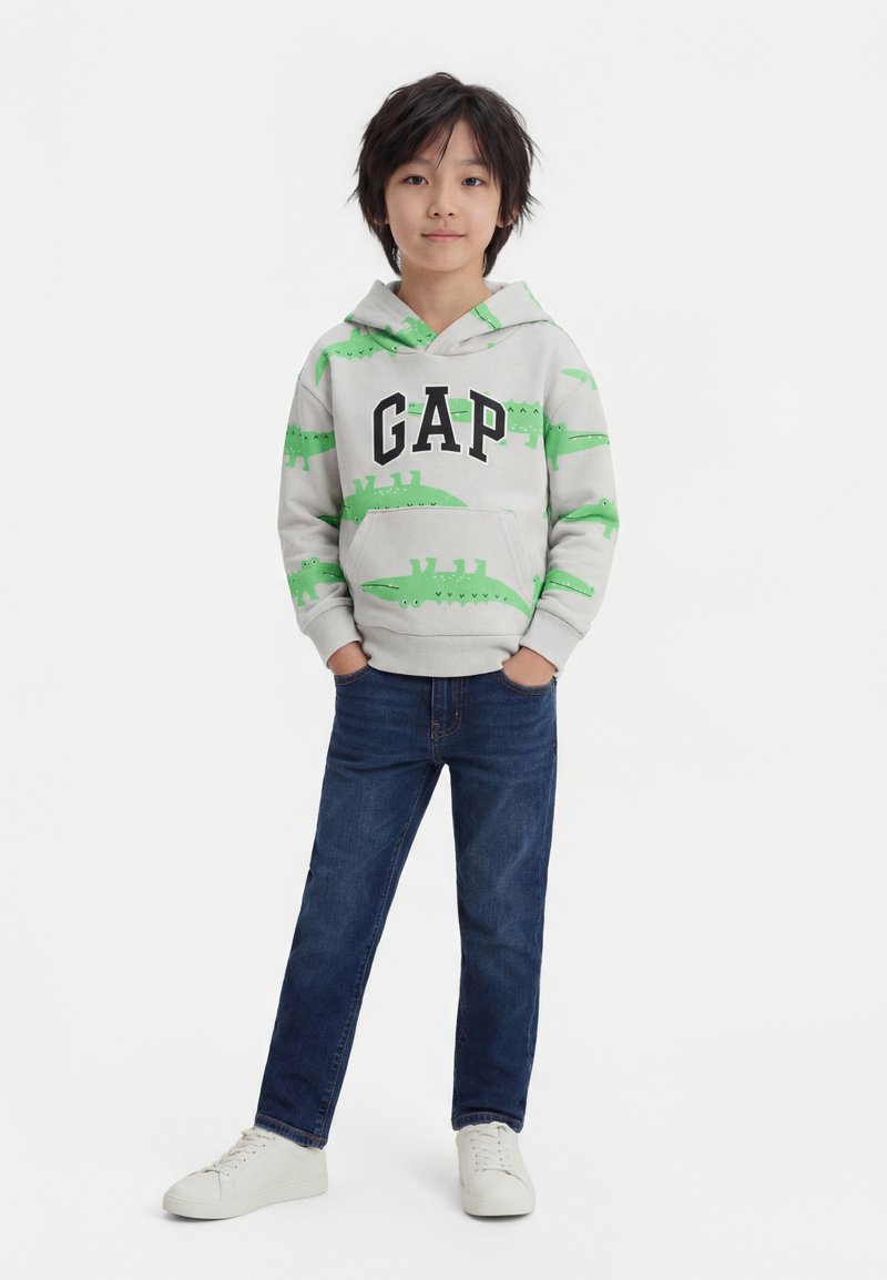 Garçon portant un sweat à capuche gris clair avec des imprimés de crocodile verts et le logo "GAP", jean bleu et baskets blanches, debout les mains dans les poches.