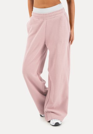 Osoba ubrana w luźne, jasnoróżowe spodnie dresowe z elastycznym białym pasem oraz białe trampki, stojąca z rękami w kieszeniach.