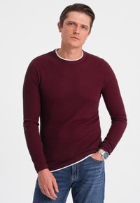 Burgundy long-sleeve shirt with a textured design and a white crew neckline, paired with blue jeans. Silver watch accents detail.