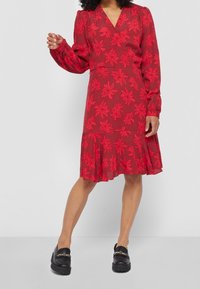 Robe florale rouge avec de longues manches bouffantes, décolleté en V et ourlet festonné. Présente des motifs de fleurs en relief. Assortie à des mocassins noirs.