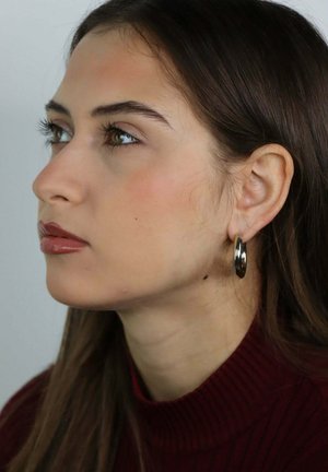 Jonge vrouw met bruin haar, gekleed in een kastanjebruine coltrui en een zilveren creooloorbel, kijkt bedachtzaam naar links tegen een neutrale achtergrond.