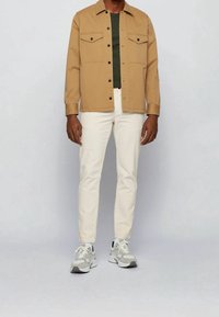 Tan knäppt jacka med två bröstfickor över en mörkgrön skjorta. Ljus beige byxor och grå sneakers fulländar outfiten.