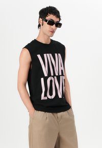 Svart ermeløs t-skjorte med fet lys rosa "VIVA LOVE"-tekst, kombinert med beige bukser og mørke rektangulære solbriller, mot en hvit bakgrunn.