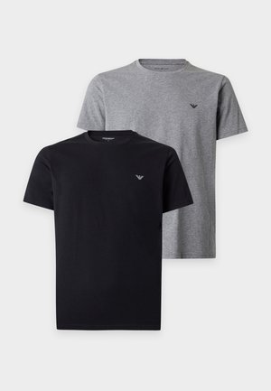 Mann steht barfuß, trägt ein schwarzes Kurzarmhemd und schwarze Hose, die Hände in den Taschen, vor einfarbigem weißem Hintergrund.