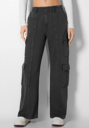 Pantalon cargo large noir à taille élastique et poches à rabat, porté avec des baskets blanches et un haut court à manches longues.
