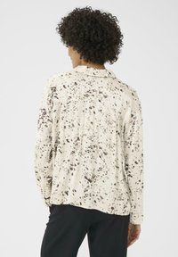 Een persoon met krullend haar is van achteren te zien, gekleed in een gespikkeld beige shirt en zwarte broek, tegen een plain witte achtergrond.