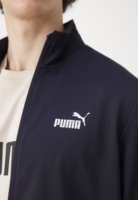 Jūras spēku zipsvārku ar gludu audumu, kuram ir balts Puma logo kreisajā krūtīs un augstas apkakles dizains.