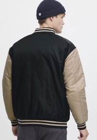 Black and tan bomber jacket with contrasting sleeves, ribbed collar and cuffs, smooth texture, and decorative stripes at the hem.
